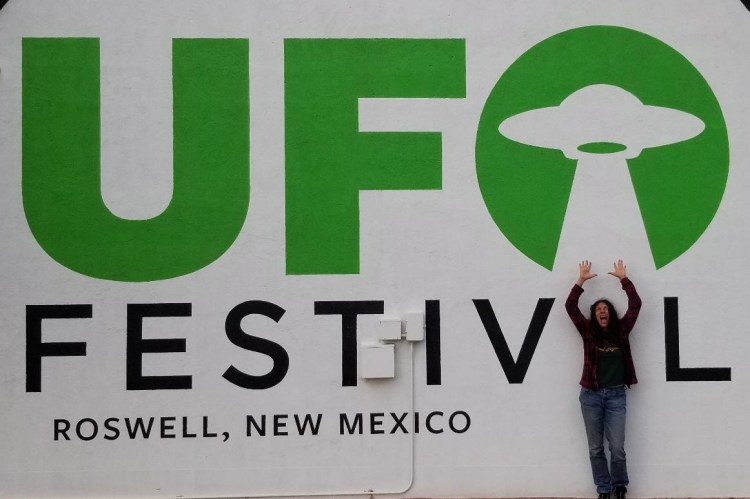 Lydia standing in front of outside of Roswell Visitor center with "UFO Festival Rowell, New Mexico"  and a UFO painted on wall